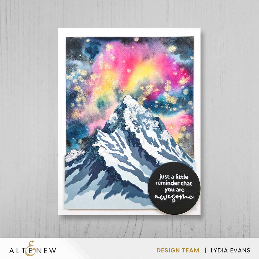 A handmade greeting card featuring a stylized snow-capped mountain under a vibrant pink, purple, yellow, and blue galaxy sky, displayed on a light gray wood-textured background. A black circular cardstock with white text is placed on the lower right of the card.  Altenew One-Go: Solid Circular Sentiments Press Plate card ideas
