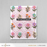 A handmade greeting card featuring a pink and white damask pattern background decorated with colorful 3D paper flowers in pink, purple, blue, and orange. A black banner in the center reads 'WE ARE SO PROUD'.