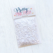 A clear plastic bag of white faux pearls, sealed with a white header card that reads 'Pretty PINK POSH' in blue and pink lettering, placed on a white wood surface. Pretty Pink Posh Marshmallow Pearls