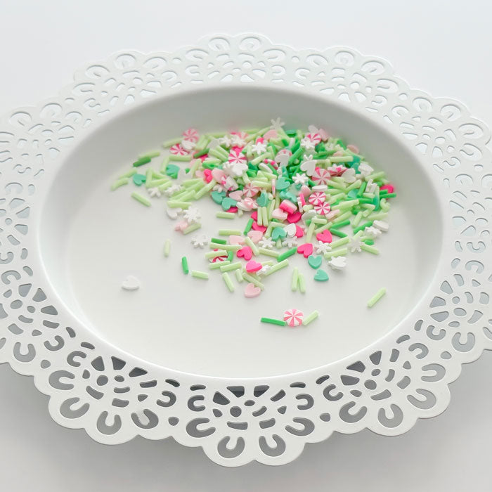 A pile of colorful sprinkles in pink, green, and white, scattered on a white ceramic plate with a decorative cutout border. Pretty Pink Posh Merry & Mint Clay Confetti