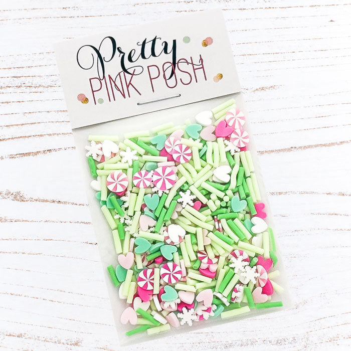 A clear plastic bag filled with pastel pink, mint green, and white decorative sprinkles and candy-shaped pieces, sealed with a white header card that reads 'Pretty Pink Posh.' The package is photographed on a white wood surface. Pretty Pink Posh Merry & Mint Clay Confetti