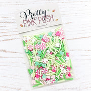A clear plastic bag filled with pastel pink, mint green, and white decorative sprinkles and candy-shaped pieces, sealed with a white header card that reads 'Pretty Pink Posh.' The package is photographed on a white wood surface. Pretty Pink Posh Merry & Mint Clay Confetti