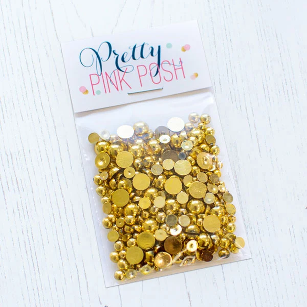 A clear plastic bag filled with round craft pearls, attached to a white header card that reads 'Pretty Pink Posh,' placed on a white wood surface. Product is called Gold Metallic Pearls.