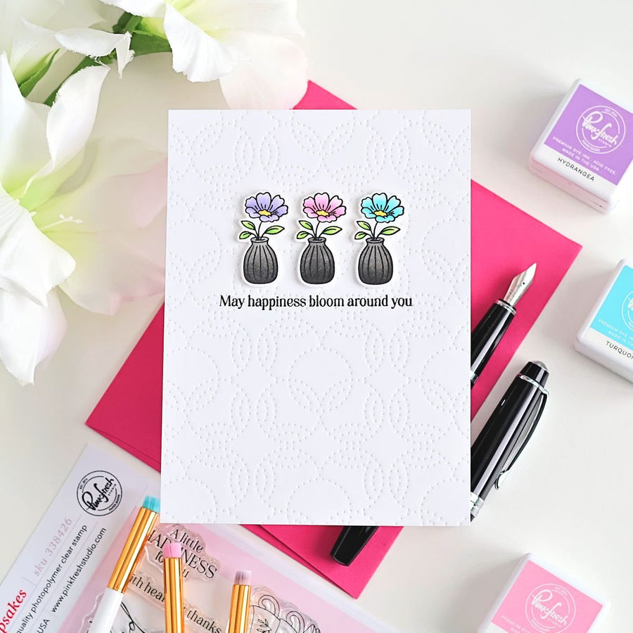 A white embossed greeting card with three colorful flower-and-bottle illustrations and the printed message 'May happiness bloom around you,' styled on a white surface with pink and magenta papers, pens, and small Ranger ink pads.