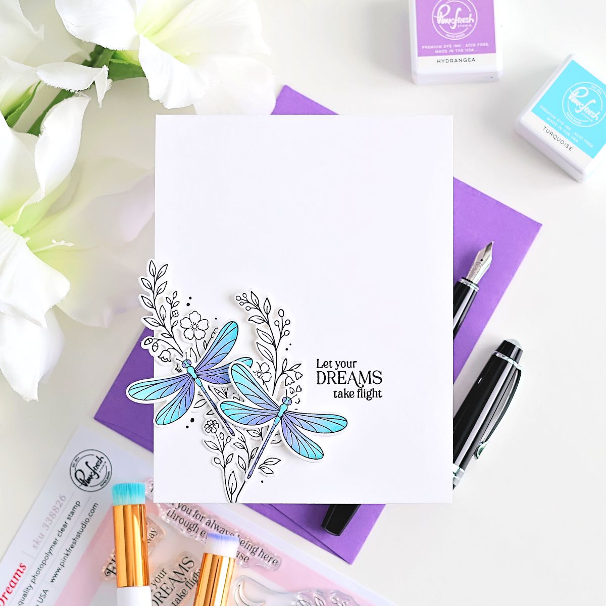 A white card featuring a hand-colored dragonfly design in blue and teal with black line art and the text 'Let your DREAMS take flight,' styled in a flat lay with purple notebooks, pens, floral props, and Ranger stamp ink pads.