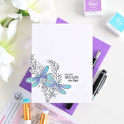 A white card featuring a hand-colored dragonfly design in blue and teal with black line art and the text 'Let your DREAMS take flight,' styled in a flat lay with purple notebooks, pens, floral props, and Ranger stamp ink pads.
