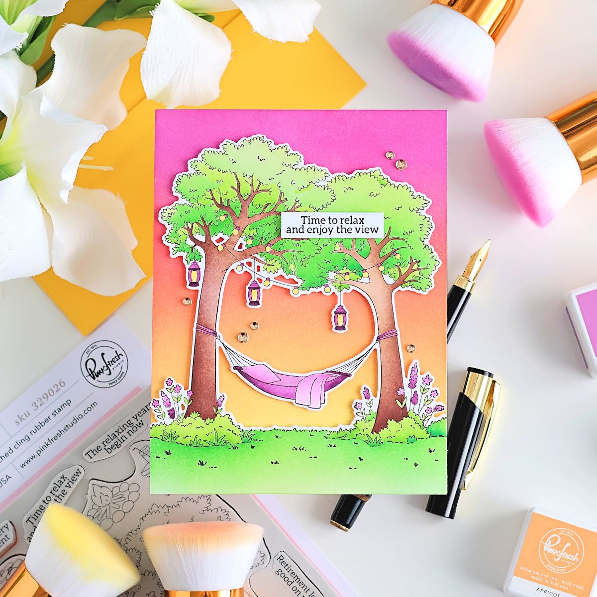A colorful handmade greeting card featuring a cartoon hammock between two trees with the text 'Time to relax and enjoy the view'. The card is styled in a flat-lay arrangement with makeup brushes, white flowers, and stamp blocks on a white surface.