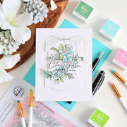 A handmade greeting card with a layered white background and a clear stamp design reading 'Sending Christmas Cheer' with blue flowers and green leaves, styled in a flat lay with crafting supplies and flowers.