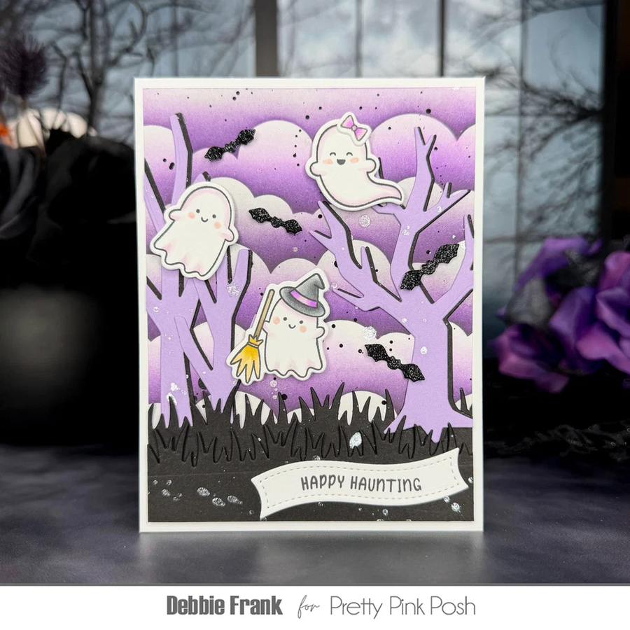 A handmade Halloween greeting card featuring cute cartoon ghosts, bats, and a broomstick among bare trees, with the text 'Happy Haunting' at the bottom. The card is displayed upright on a dark surface with a blurred window and purple flowers in the background.