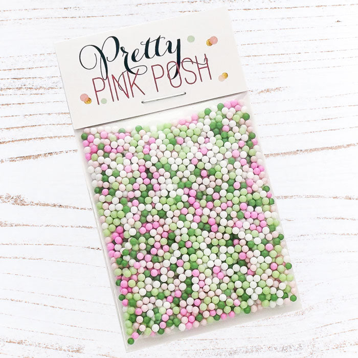 A clear plastic bag filled with pastel pink, green, and white round craft sprinkles, sealed with a white header card printed with the brand name 'Pretty Pink Posh' on a white wood-textured background. Pretty Pink Posh Mistletoe Kisses Shaker Beads