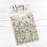 A clear plastic bag filled with pastel pink, green, and white round craft sprinkles, sealed with a white header card printed with the brand name 'Pretty Pink Posh' on a white wood-textured background. Pretty Pink Posh Mistletoe Kisses Shaker Beads