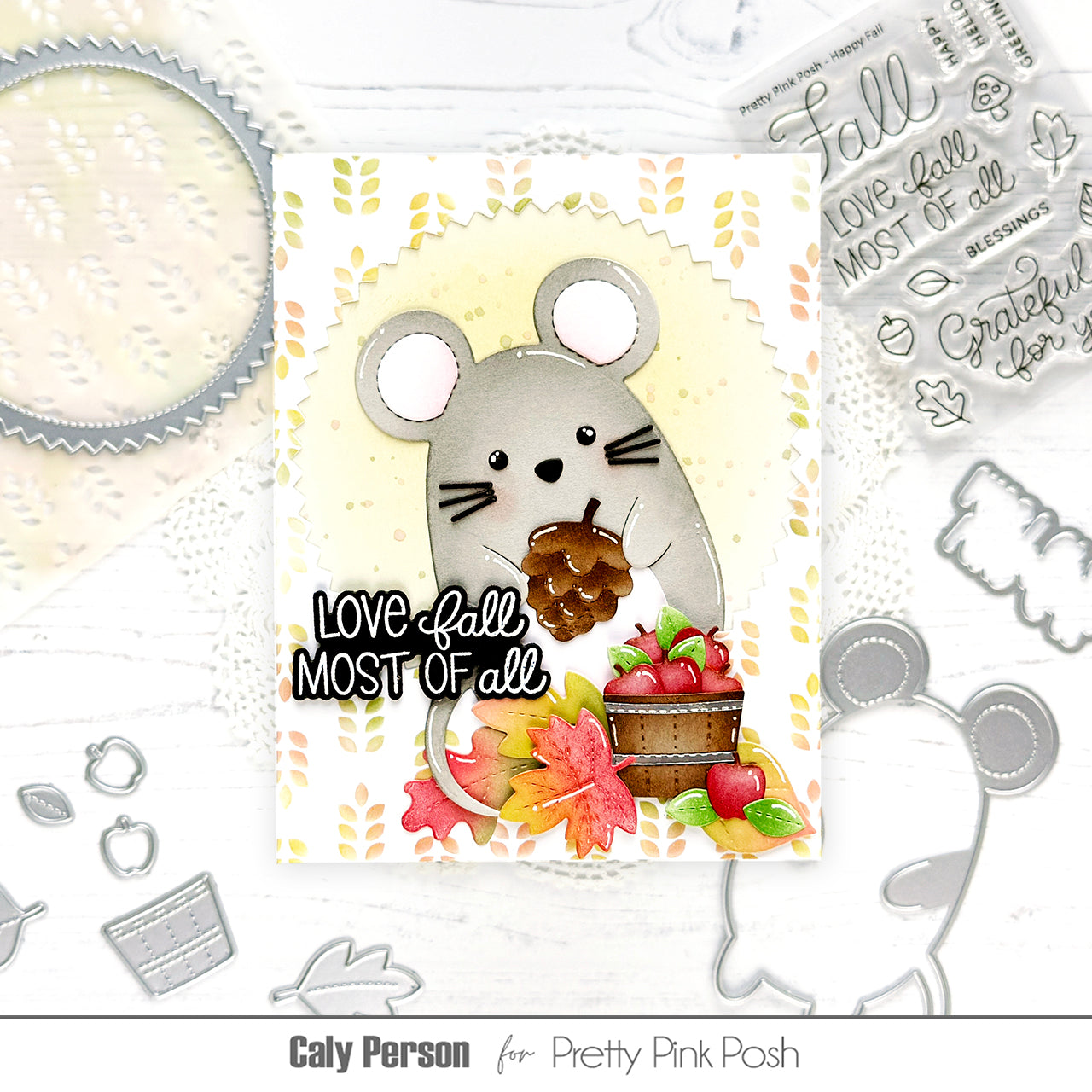 A handmade mouse card featuring a gray mouse holding a pinecone, with autumn leaves, apples, and the phrase 'Love fall most of all' on a white card. The card is displayed on a white surface surrounded by crafting tools and clear stamps, with text reading 'Caly Person for Pretty Pink Posh'.