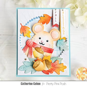 A handmade autumn-themed greeting card featuring a cute mouse character wearing a red scarf, surrounded by colorful fall leaves and acorns. The card is presented on a white wooden surface with decorative flowers in the background.