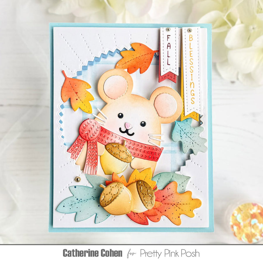 A handmade autumn-themed greeting card featuring a cute mouse character wearing a red scarf, surrounded by colorful fall leaves and acorns. The card is presented on a white wooden surface with decorative flowers in the background.
