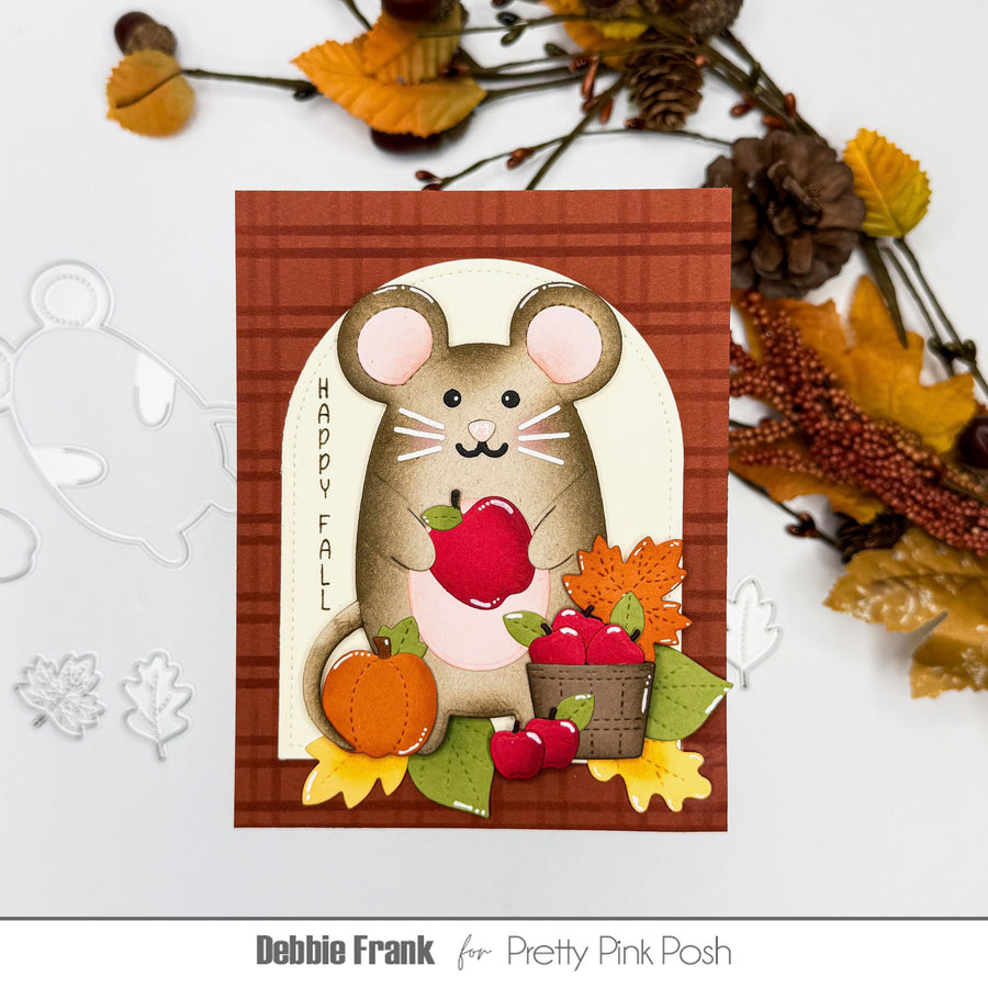 A handmade autumn-themed card featuring a mouse holding a red apple, surrounded by fall leaves, pinecones, and a basket of apples. The card is displayed on a white surface with decorative autumn leaves and pinecones around it.