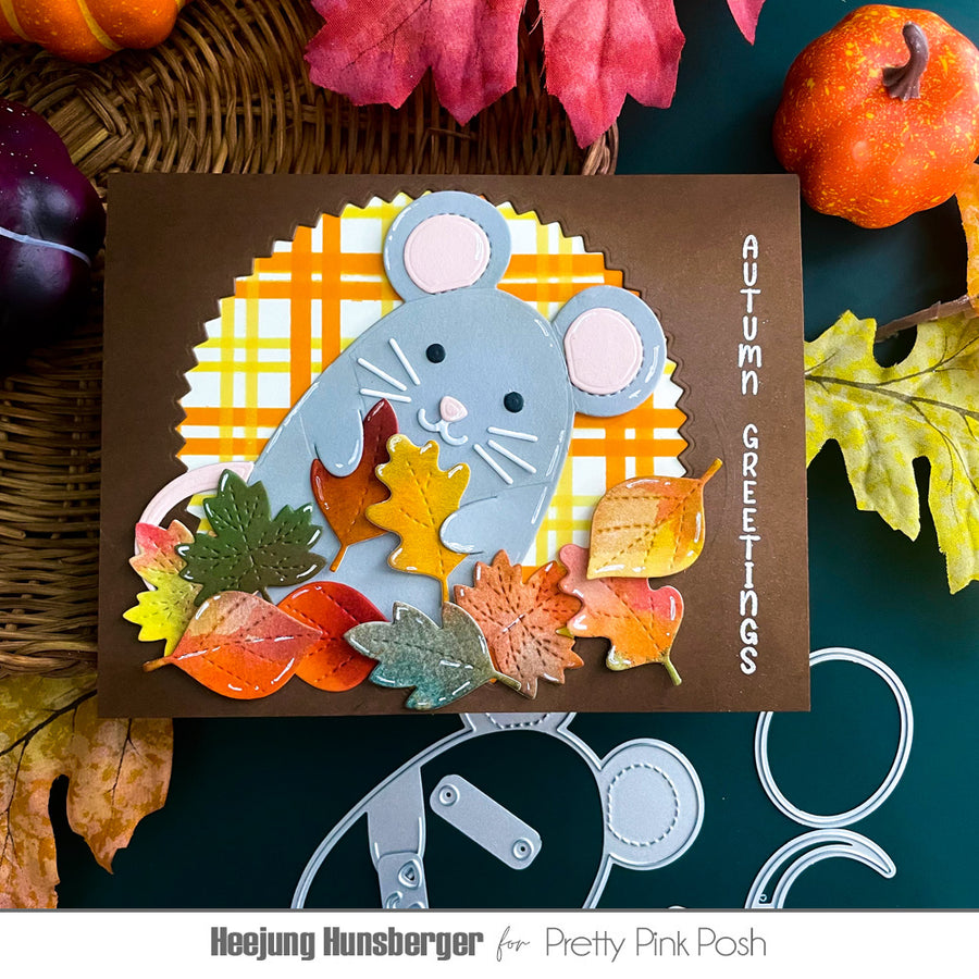 A handmade autumn-themed greeting card featuring a light blue mouse, colorful layered leaves, and a yellow-orange plaid circle on a brown background. The card is displayed in a flat-lay arrangement with pumpkins, fall leaves, and crafting tools.