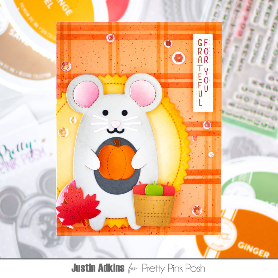 A handmade greeting card featuring a white mouse holding a pumpkin, with autumn-themed details like a red maple leaf, a basket of apples, and a plaid orange background. The card is displayed on a styled flat-lay background with other cards and stamps.