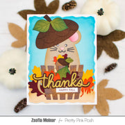 A handmade autumn-themed greeting card featuring a cute beige hamster wearing a brown acorn hat, standing behind a wooden fence with colorful fall leaves and the words 'thanks' and 'HAPPY FALL'. The card is displayed on a white wooden surface with decorative pumpkins, leaves, and pinecones around it.