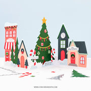 A festive pop-up cardboard Christmas village scene with a decorated green tree, pink and red houses, candy canes, and small paper trees on a white snowy base against a pale blue background. A website URL is printed at the bottom.