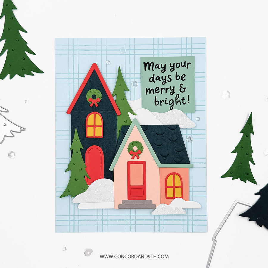 A handmade layered paper holiday card featuring two cozy houses, snow, pine trees, and a green message box that reads 'May your days be merry & bright!'. The card is displayed on a white background with subtle decorative paper cutouts around it.