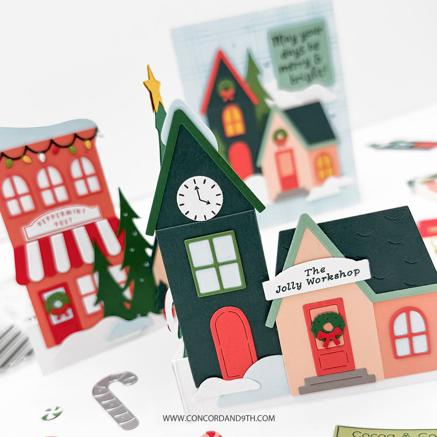 A close-up of a handmade paper pop-up holiday village scene with layered house and tree cutouts. The main pop-up is a dark green house labeled "The Jolly Workshop" with a white sign, red door, peach walls, and green roof.