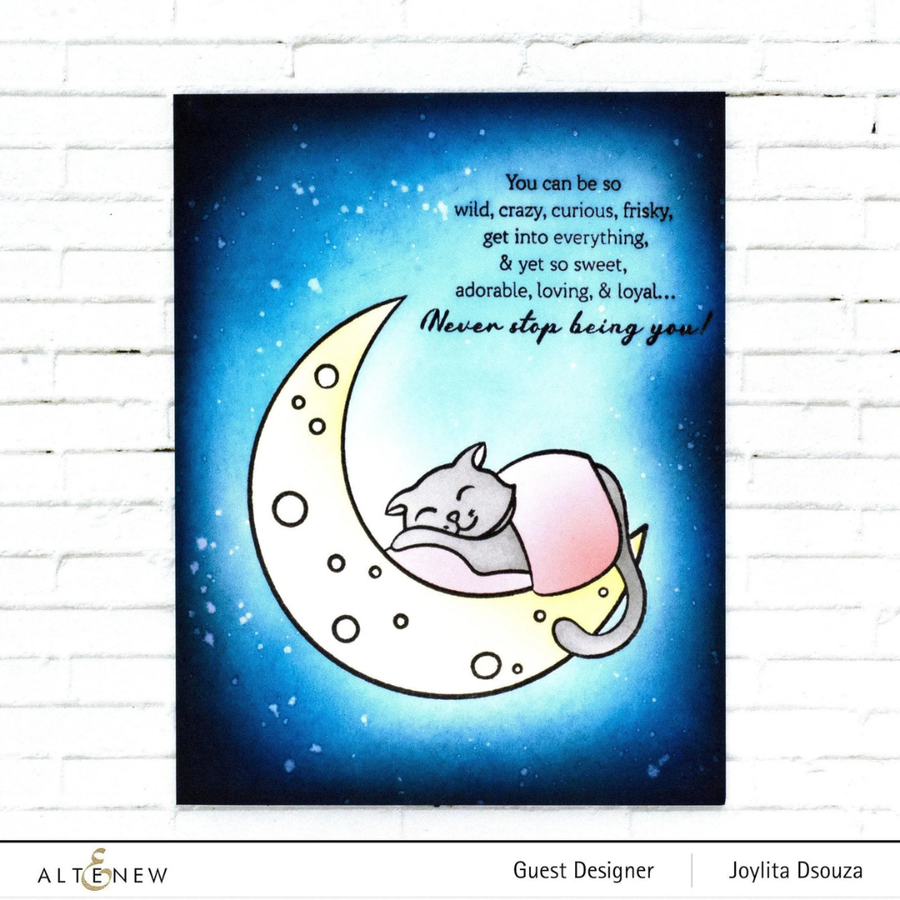 A handmade greeting card featuring a sleeping gray cat curled up inside a pale yellow crescent moon on a starry blue night sky. White script text appears near the top right, and the print is shown hanging against a white brick wall. Black print stamped with Altenew Obsidian Pigment Ink