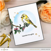 A handmade greeting card featuring a yellow bird perched on a branch with pink berries against a light blue circle, with the text 'heartfelt thoughts' at the bottom. The card is styled on a light surface with peach flowers, gold scissors, and small gold embellishments.
