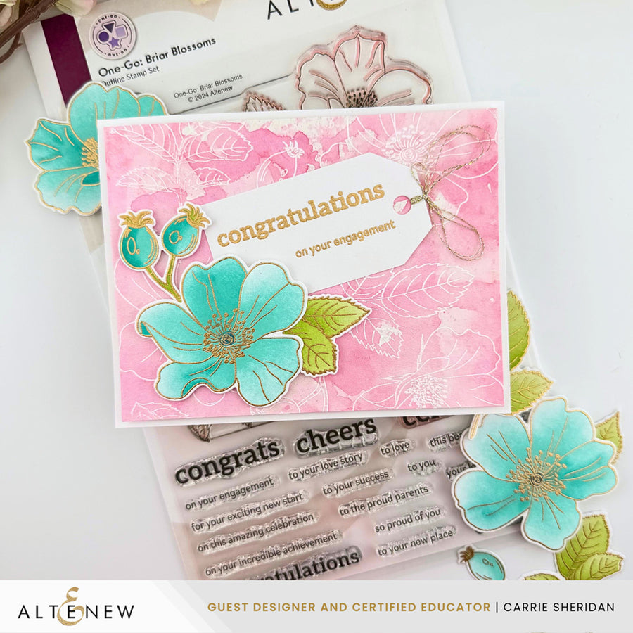 A handmade engagement congratulations card featuring a pink watercolor background with a large turquoise flower, green leaves, and a white tag reading 'congratulations on your engagement.' The card is styled on top of other floral embellishments, stamps, and cards from Altenew. Altenew One-Go: Briar Blossoms card ideas
