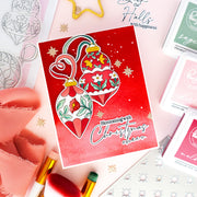 A red Christmas card featuring two hanging ornament illustrations with floral details and the text 'Blossoming with Christmas cheer,' styled on a pink surface with ribbon, stamps, and small decorative items.