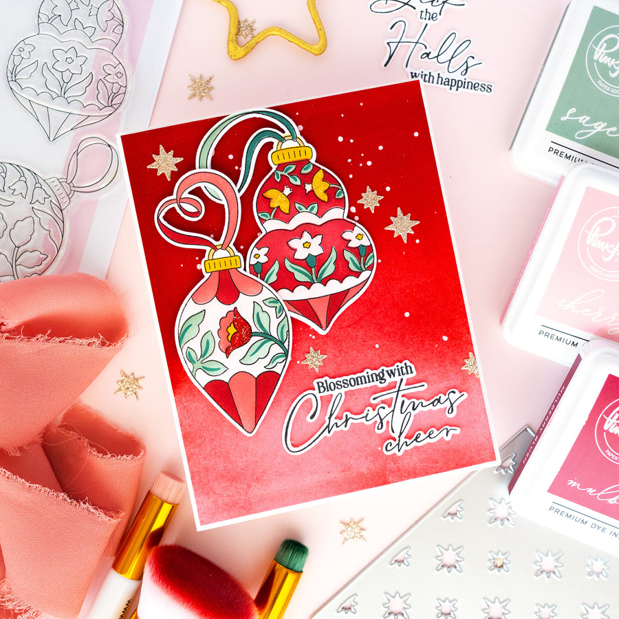 A red Christmas card featuring two hanging ornament illustrations with floral details and the text 'Blossoming with Christmas cheer,' styled on a pink surface with ribbon, stamps, and small decorative items.