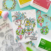 A handcrafted Christmas card featuring two colorful ornament illustrations with floral details and the text 'Warmest wishes this Christmas,' styled on a mint background with dye ink pads, stamps, and brushes.