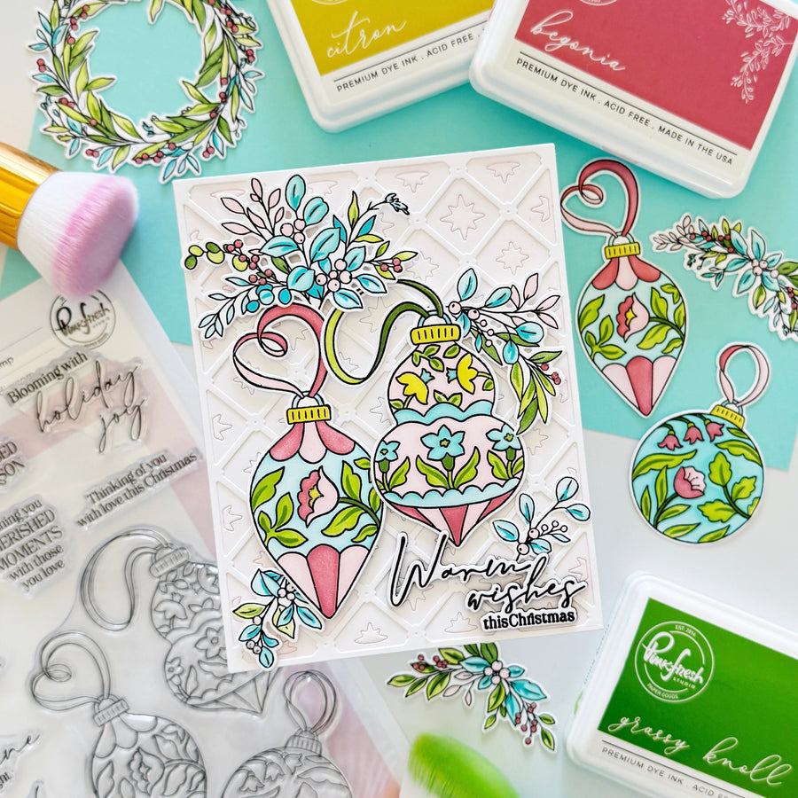 A handcrafted Christmas card featuring two colorful ornament illustrations with floral details and the text 'Warmest wishes this Christmas,' styled on a mint background with dye ink pads, stamps, and brushes.