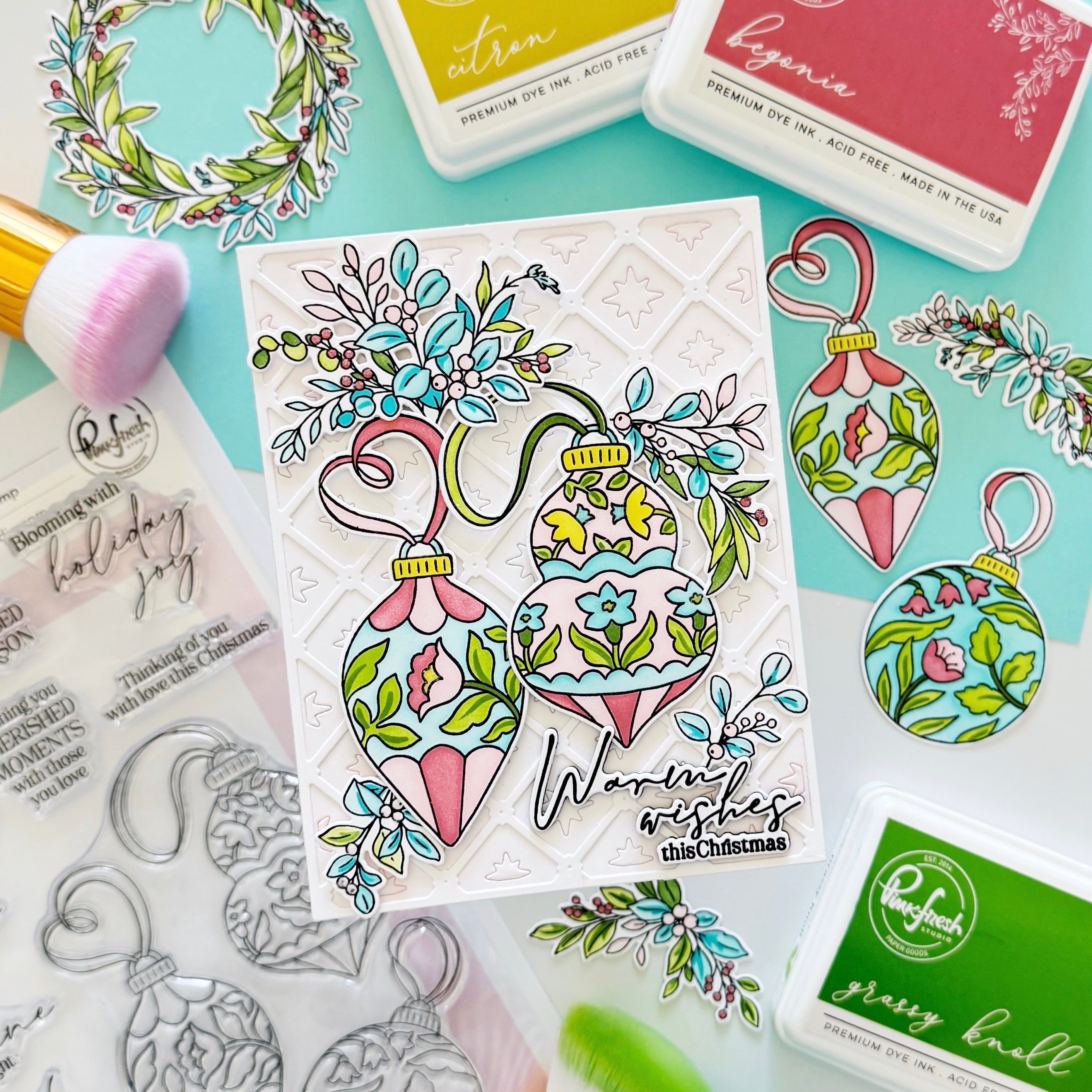 A handcrafted Christmas card featuring two colorful ornament illustrations with floral details and the text 'Warmest wishes this Christmas,' styled on a mint background with dye ink pads, stamps, and brushes.