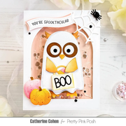 A handmade layered Halloween card featuring a ghost owl with large brown eyes, a yellow bow tie, and a 'BOO' sign, set against a peach arch with confetti, a spider on a web, and small pumpkins. The card is photographed on a white wood surface with decorative flowers in the corners.