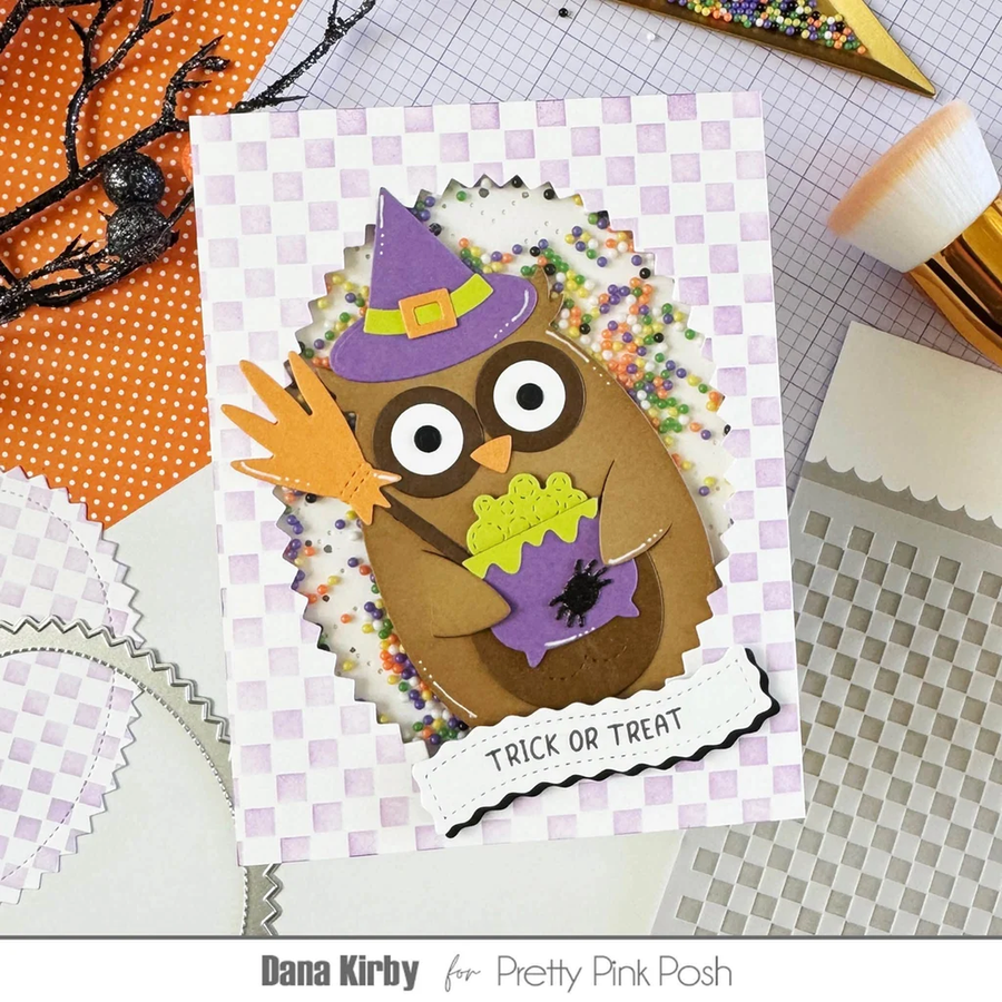 A handmade Halloween greeting card featuring a brown owl wearing a purple witch hat and holding a purple spider, surrounded by colorful confetti and a lavender checkered border. The card is displayed on a craft mat with other paper crafting tools and materials.
