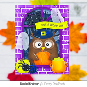 A Halloween-themed handmade card or featuring a brown owl in a witch hat holding an orange pumpkin, with a purple brick-patterned border, ghost decorations, a black spider, a green cauldron, and a yellow banner reading 'HAVE A SPOOKY DAY.' The image is presented on a white background with blurred autumn leaves around it.