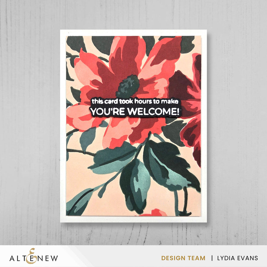 A framed floral greeting card with large red and pink flowers, dark green leaves, and the text 'this card took hours to make YOU'RE WELCOME!' centered on a light gray wood background. The Altenew logo and 'Design Team | Lydia Evans' text appear along the bottom.