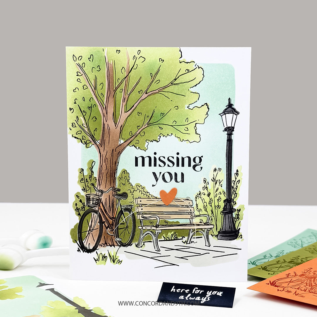 A handmade greeting card featuring a watercolor-style illustration of a tree, bicycle, bench, and lamppost with the text 'missing you' and 'here for you always'. The card is displayed upright on a white surface with other colorful card pieces and stamps nearby.