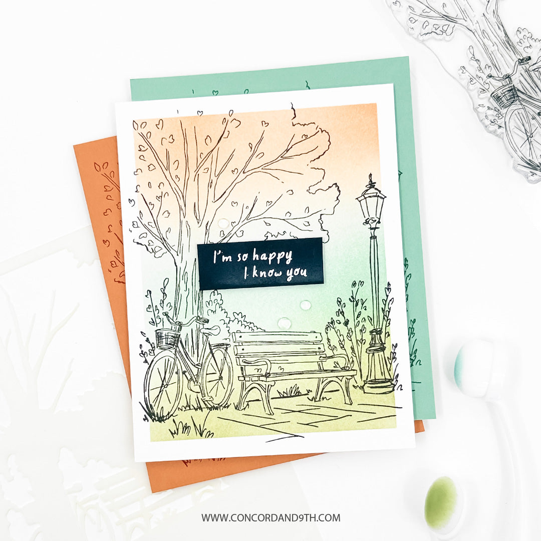 A handmade greeting card featuring a hand-drawn illustration of a bicycle, bench, and lamppost under a pastel sunset sky, with the text 'I'm so happy I know you'. The card is layered over orange and mint green cardstock and photographed on a white surface with decorative elements.