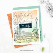A handmade greeting card featuring a hand-drawn illustration of a bicycle, bench, and lamppost under a pastel sunset sky, with the text 'I'm so happy I know you'. The card is layered over orange and mint green cardstock and photographed on a white surface with decorative elements.