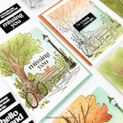A close-up of several handmade greeting cards arranged on a white surface. The cards feature illustrated trees, benches, bicycles, and street lamps in a watercolor-style autumn palette, with black text stickers reading phrases like 'missing you' and 'I'm so happy to see you.