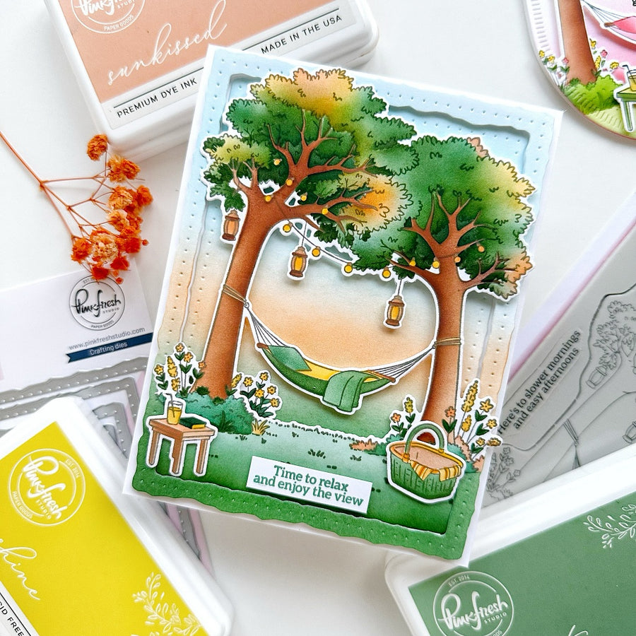 A handmade layered card featuring a camping scene with two trees, a hammock, lanterns, and the text 'Time to relax and enjoy the view', styled on top of various craft ink pads and stamp papers.