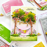 A colorful handmade greeting card featuring two illustrated trees with green, yellow, and pink foliage, a hammock between them, and the text 'Time to relax and enjoy the view.' The card is styled on a flat-lay with Ranger brand craft supplies, including an ink pad, stamping plates, and a small flower.