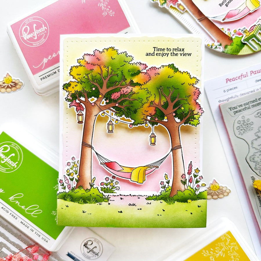 A colorful handmade greeting card featuring two illustrated trees with green, yellow, and pink foliage, a hammock between them, and the text 'Time to relax and enjoy the view.' The card is styled on a flat-lay with Ranger brand craft supplies, including an ink pad, stamping plates, and a small flower.