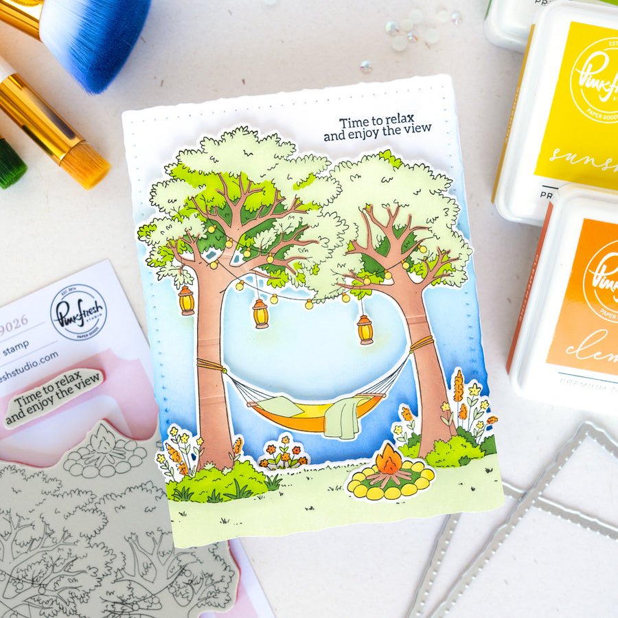 A handmade card featuring a whimsical camping scene with two trees, a hammock, a campfire, and the text 'Time to relax and enjoy the view.' The card is displayed on a light surface with crafting supplies and ink pads around it.