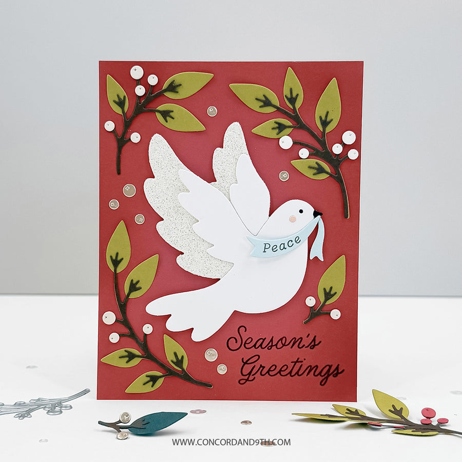 A handmade layered greeting card with a white dove carrying a banner that says 'Peace' and the words 'Season's Greetings' in black script. The card has a red background, green and gold leafy branches, and small white berries, photographed on a white surface with a light gray background.