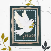 A handmade layered greeting card featuring a white dove holding an olive branch, with the word 'peace' in metallic script at the bottom. The card has a dark teal background, a beige inner border, and a light blue scalloped inner frame, styled on a white background with decorative leaf cutouts.