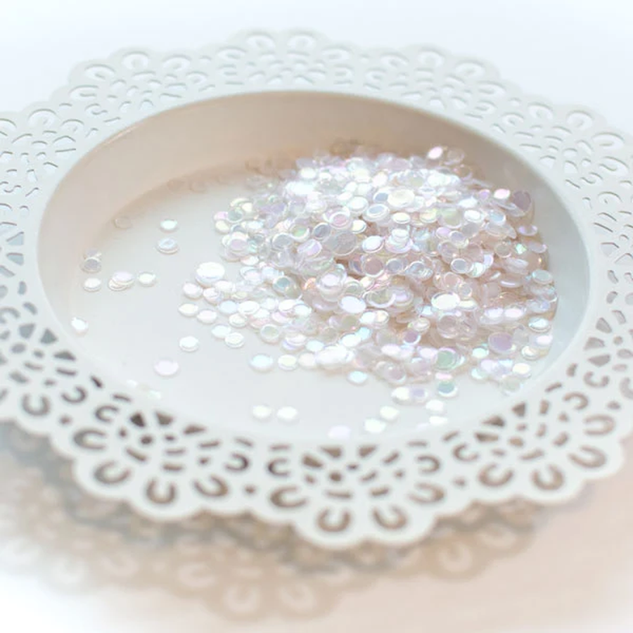 A pile of small round iridescent discs in a white ceramic dish with a scalloped edge, photographed on a soft white background. Pretty Pink Posh Pearl Prism Confetti Mix