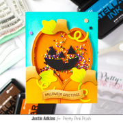 A handmade Halloween greeting card featuring a layered orange jack-o'-lantern with yellow leaves, a black stitched face, multicolored sprinkles, and a tan banner reading 'Halloween Greetings' on a turquoise background. The card is photographed on a white surface with craft supplies and an Ink Pad visible around it.