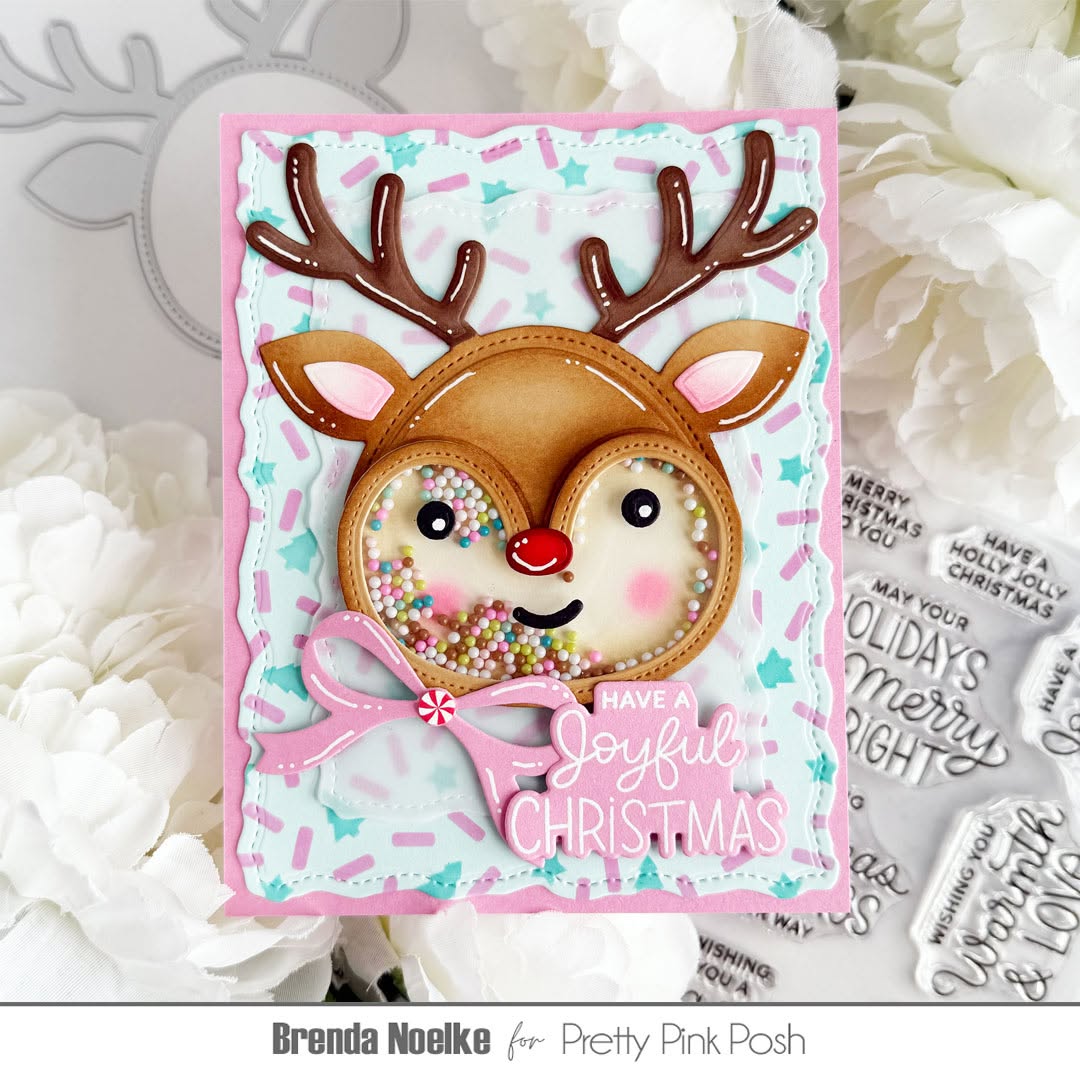 A handmade Christmas greeting card featuring a reindeer face with antlers, a pink bow, and the phrase 'Have a Joyful Christmas' on a pastel star-patterned background, styled with white flowers and holiday-themed stamps.